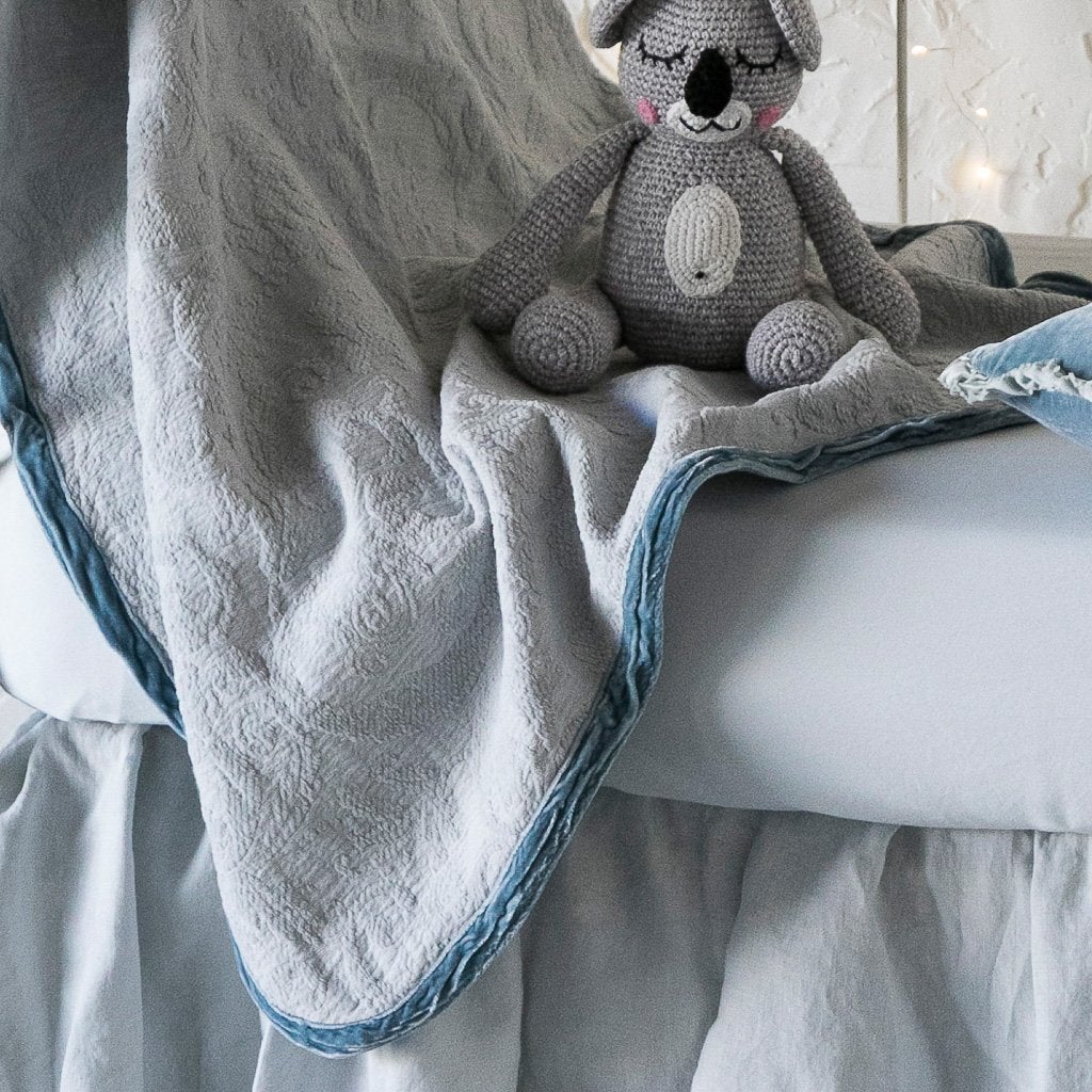 : A close up of the Adele baby blanket in cloud draped over a crib mattress, highlighting the texture of the cotton damask and the silk velvet edging. The trim absorbs the dye more richly than the cotton body.