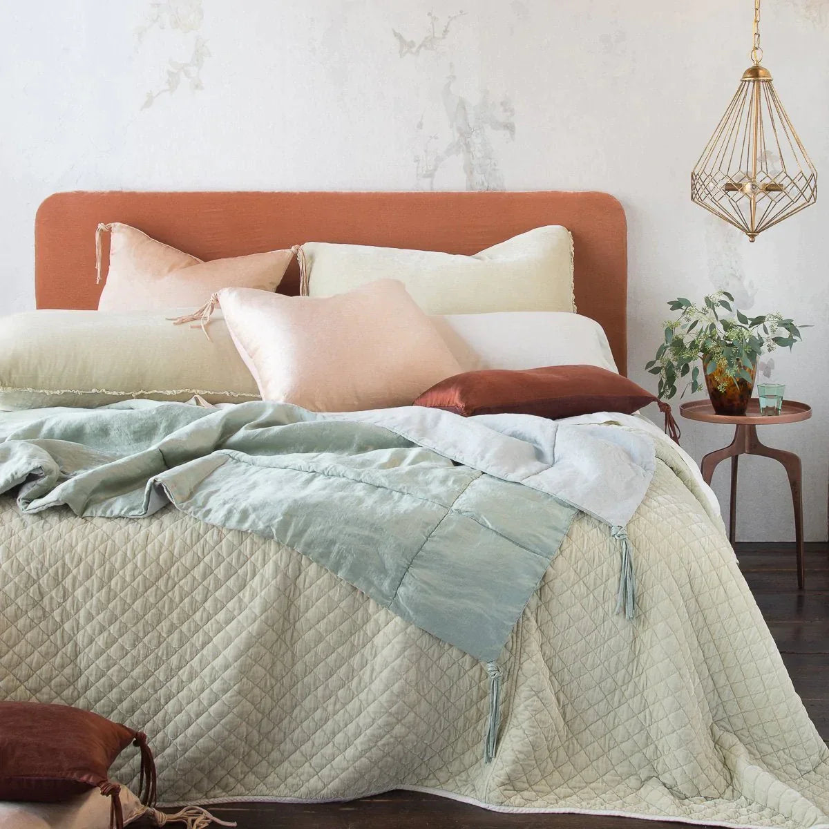: Carmen shams on a partially unmade bed, with silk velvet and silk charmeuse accessory pices in creams, blues, pinks, and browns - end of bed view.