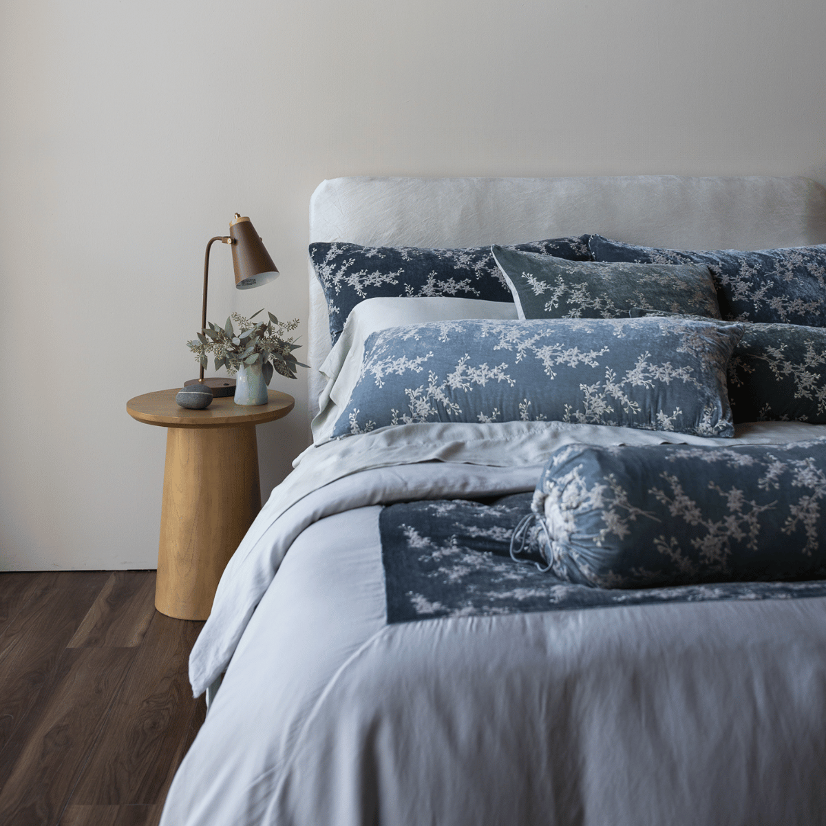 : a grey-ish blue duvet cover with several matching blue and green toned pillows against a white charmeuse headboard and medium wood side table.