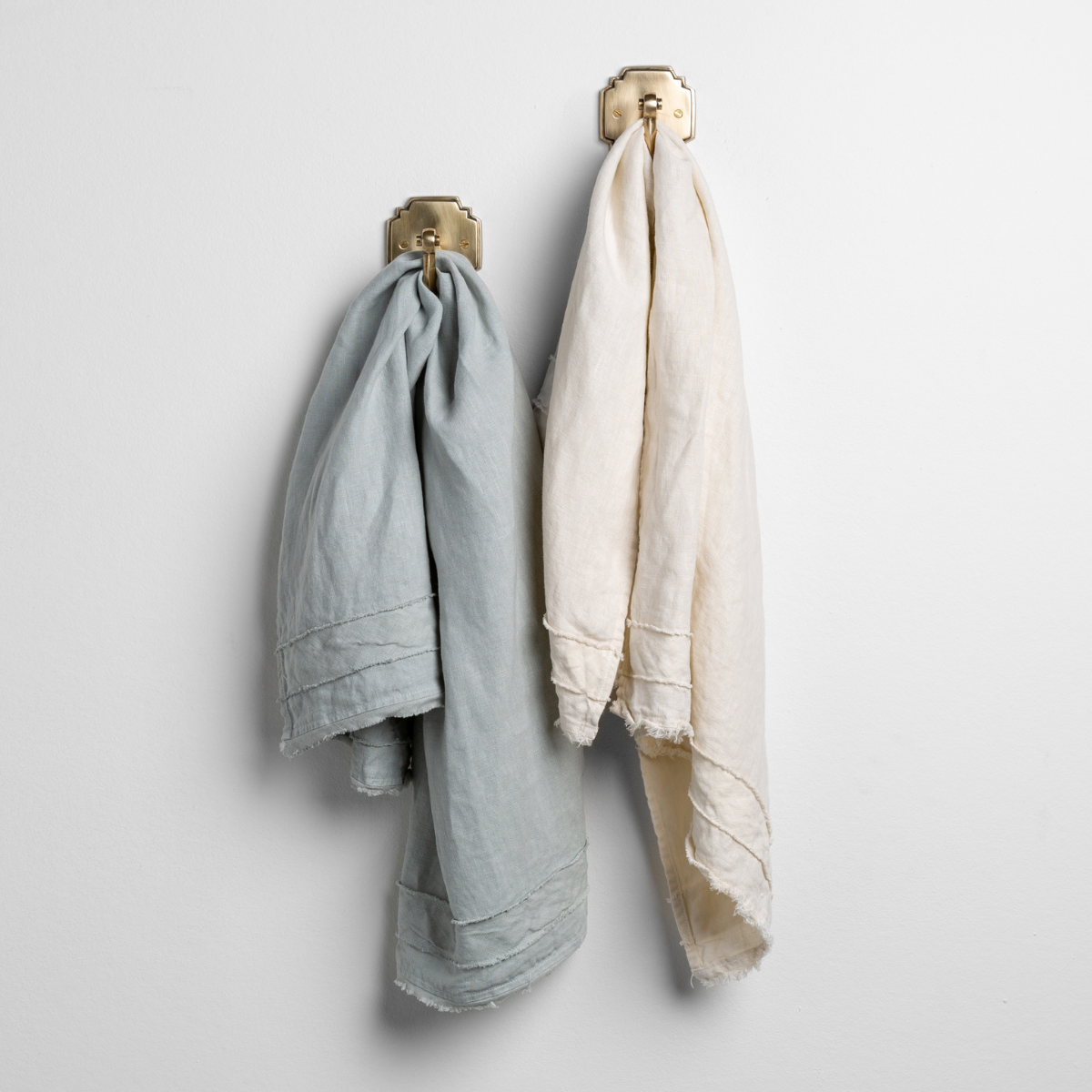 : a pair of midweight linen guest towels in cloud and parchment on gold towel hooks mounted to a white wall.