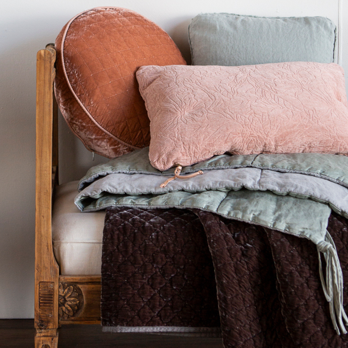 : A close up of one side of a daybed bench styled with throw pillows and throw blankets in grey, light green and warm tones.