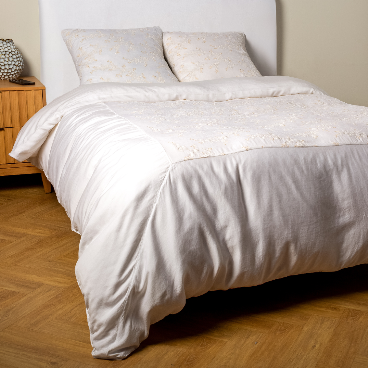 White: A ¾ angle shot of an embroidered silk velvet and tencel duvet cover and matching shams with a vase and cell phone on the side table.