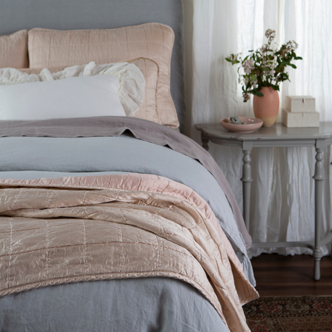 : shot from the foot of the bed, a quilted charmeuse blanket is in the foreground and the shams and decorative throw pillows slightly out of focus at the head of the bed.