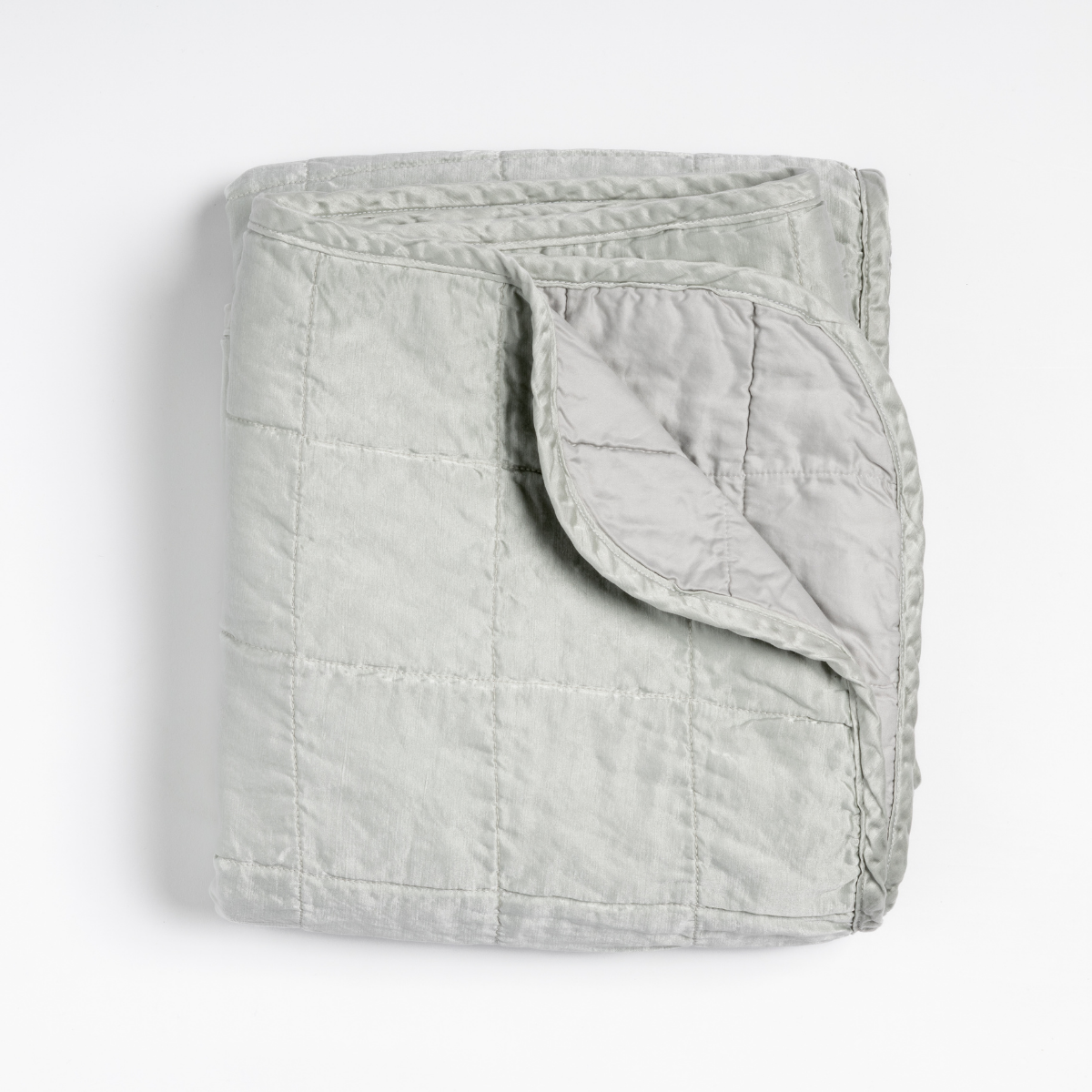 Cloud: an overhead shot of a quilted charmeuse blanket in cloud, a subtle sky blue-grey, with a corner folded down, showing its cotton sateen back.