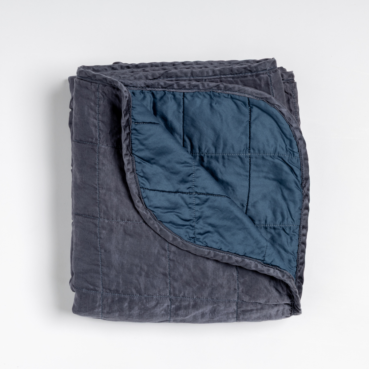 Midnight: an overhead shot of a quilted charmeuse blanket in midnight, a rich indigo tone, with a corner folded down, showing its cotton sateen back.