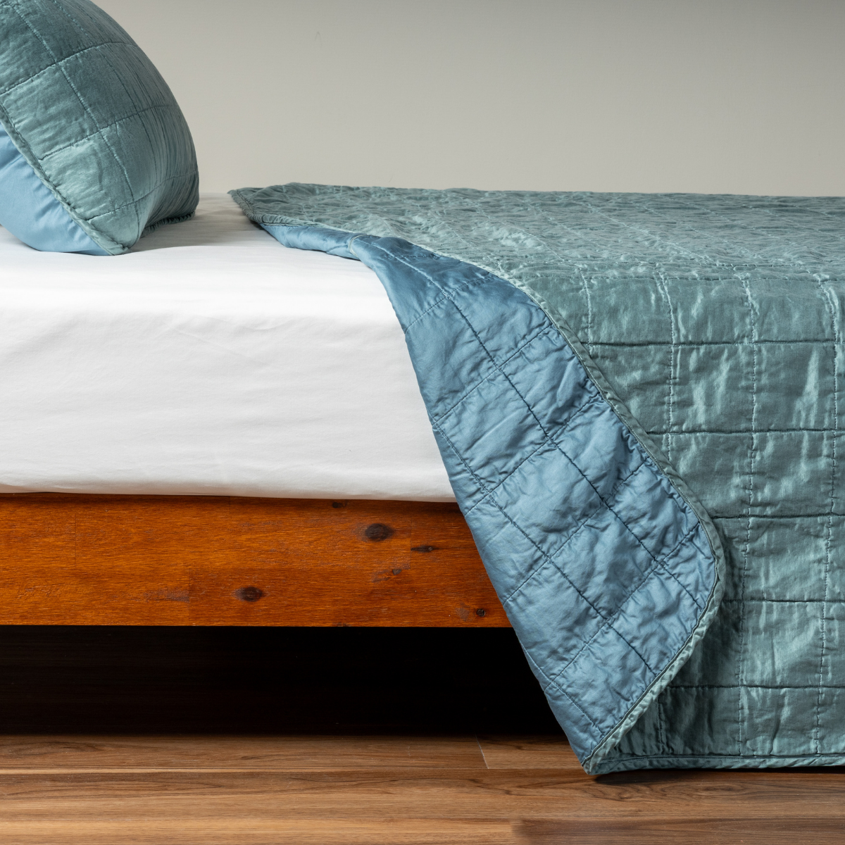 Cenote: A side shot of a square quilted charmeuse coverlet in cenote, a vibrant ocean-inspired blue-green, with its matching sham, the corner of the coverlet peeled back, revealing the cotton sateen back.