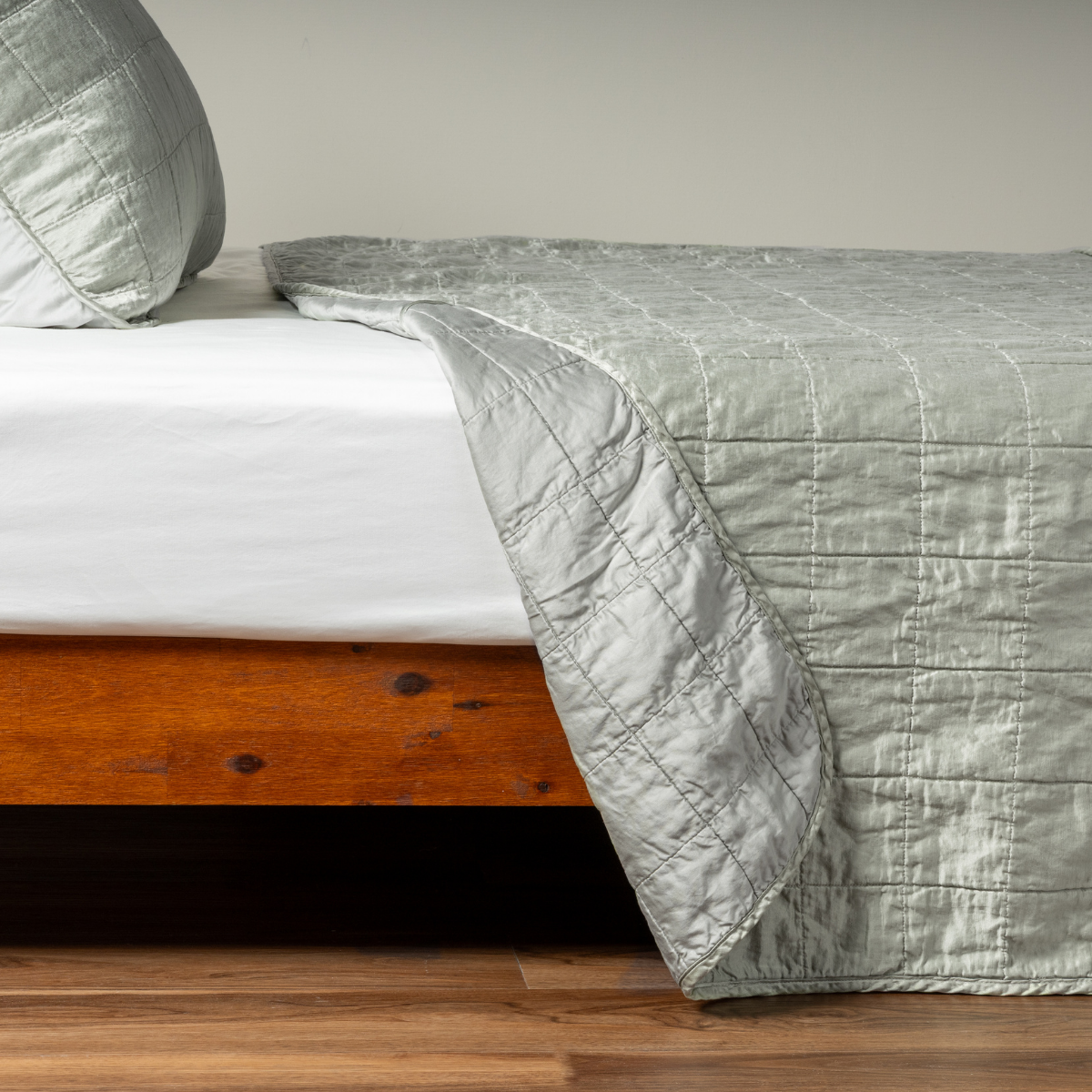 Eucalyptus: A side shot of a square quilted charmeuse coverlet in eucalyptus, a soft light green, with its matching sham, the corner of the coverlet peeled back, revealing the cotton sateen back.