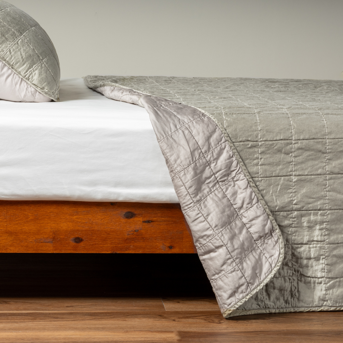 Fog: A side shot of a square quilted charmeuse coverlet in fog, a soft mid-tone grey, with its matching sham, the corner of the coverlet peeled back, revealing the cotton sateen back.