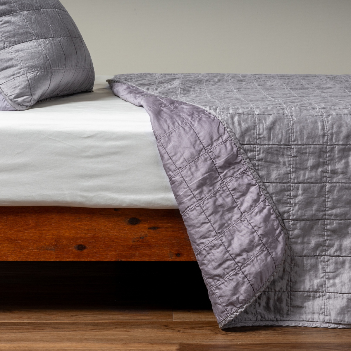 French Lavender: A side shot of a square quilted charmeuse coverlet in french lavender, a neutral violet tone, with its matching sham, the corner of the coverlet peeled back, revealing the cotton sateen back.