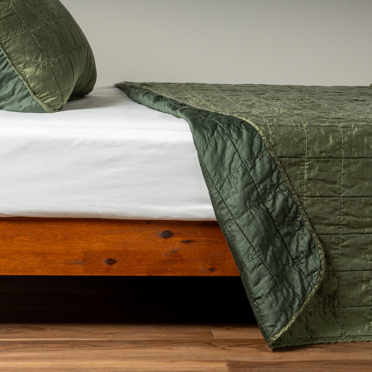 Juniper: A side shot of a square quilted charmeuse coverlet in juniper, a deeply saturated dark green, with its matching sham, the corner of the coverlet peeled back, revealing the cotton sateen back.