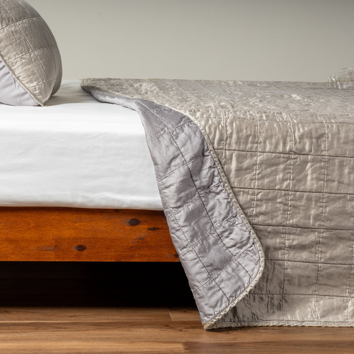 Moonlight: A side shot of a square quilted charmeuse coverlet in moonlight, a saturated, cool, mid-dark grey tone, with its matching sham, the corner of the coverlet peeled back, revealing the cotton sateen back.