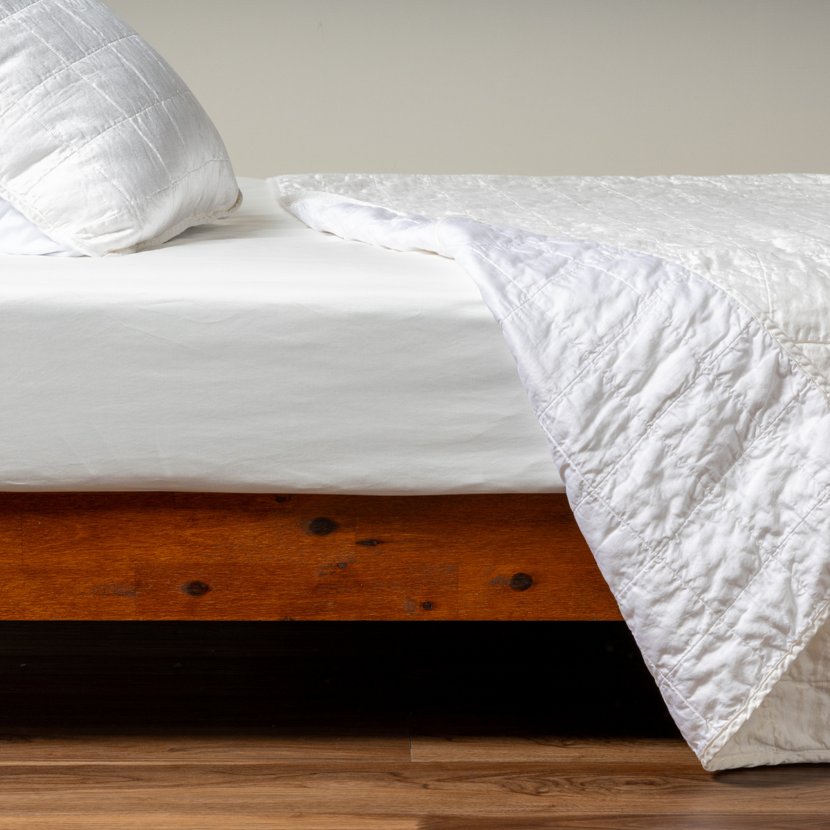 White: A side shot of a square quilted charmeuse coverlet in white, with its matching sham, the corner of the coverlet peeled back, revealing the cotton sateen back.