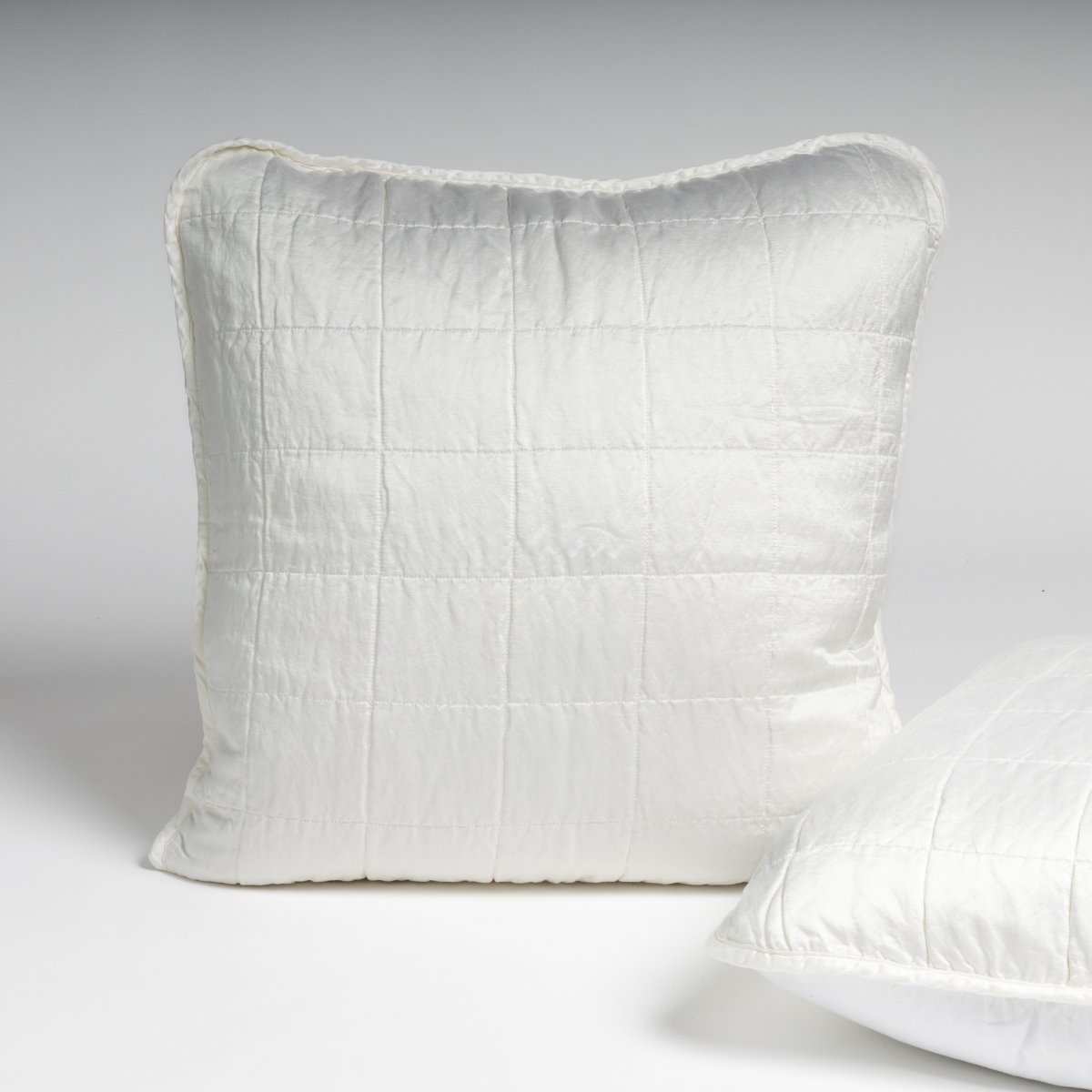 White: A pair of shams shown in white against a white background with one sham upright and the other on its side to show the cotton sateen back and square-quilted charmeuse front.