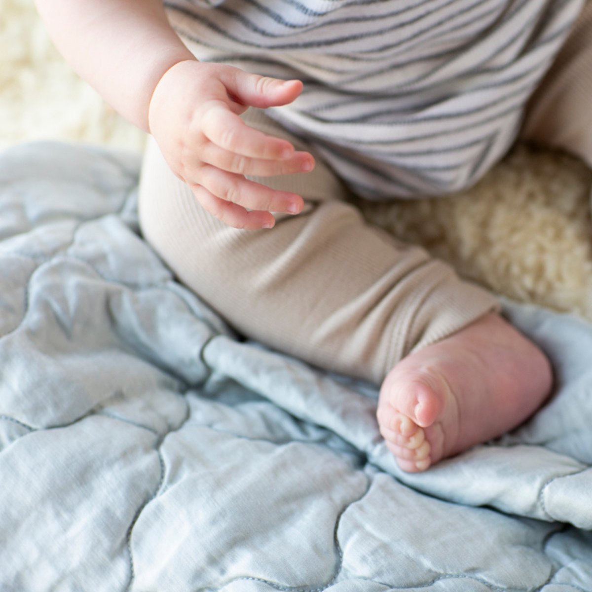 : Angled overhead close up view of rumpled blanket in cloud, shown with wooden toys and partial view of a sitting baby with bare feet.