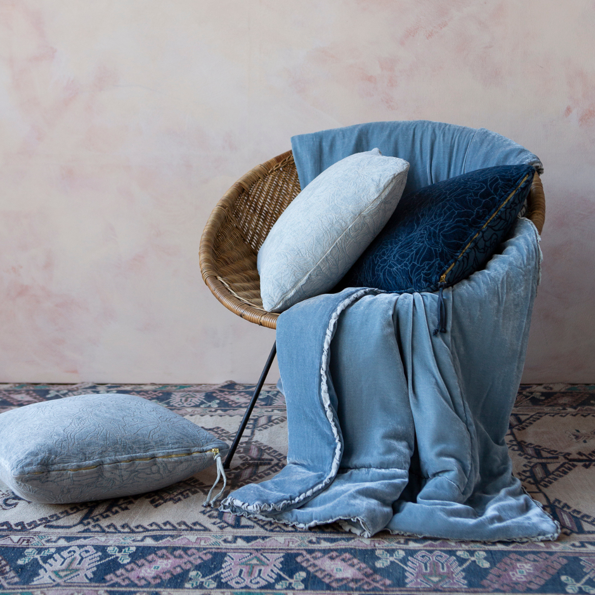 : a papasan-shaped chair is draped with a silk velvet throw blanket and coordiinating chenille and cotton jacquard throw pillows in shades of blue — shown against a pink-toned wall and colorful rug.