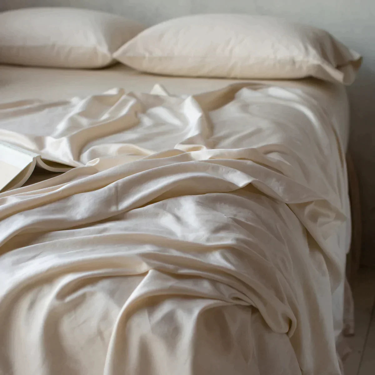 : Bria flat sheet in parchment with matching fitted sheet and standard pillows, shown from three-quarter angle. The flat sheet is rumpled, emphasizing the fabric's sheen.