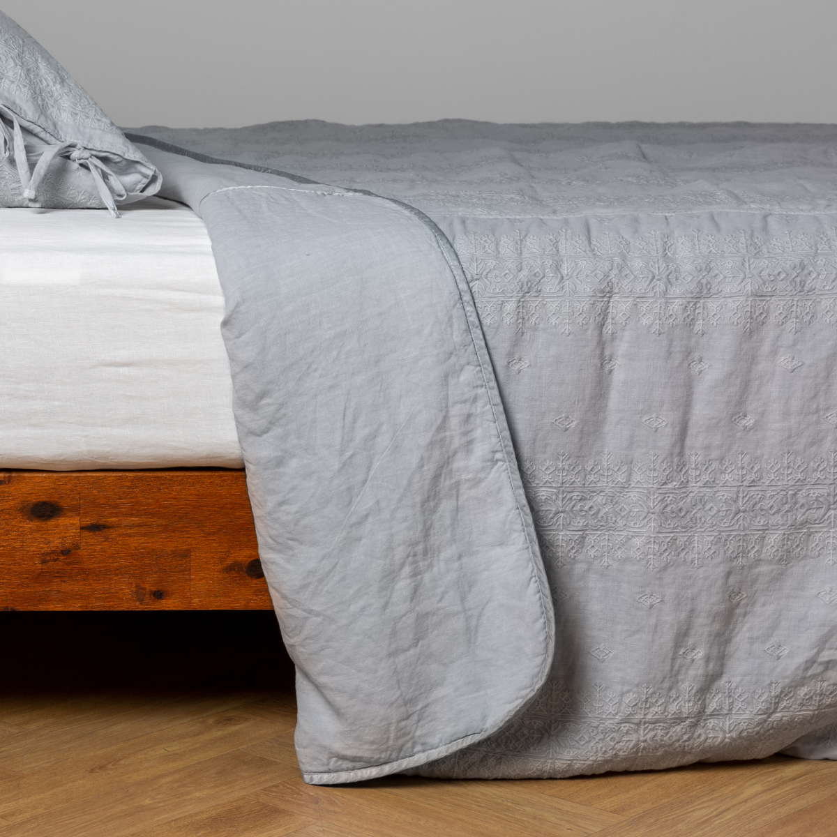 Cloud: White: embroidered bedspread with corner pulled back showing the plain linen back. Shown with a white fitted sheet and matching sham, the bedframe is slightly visible and the bed is against a white wall and on a medium wood floor.