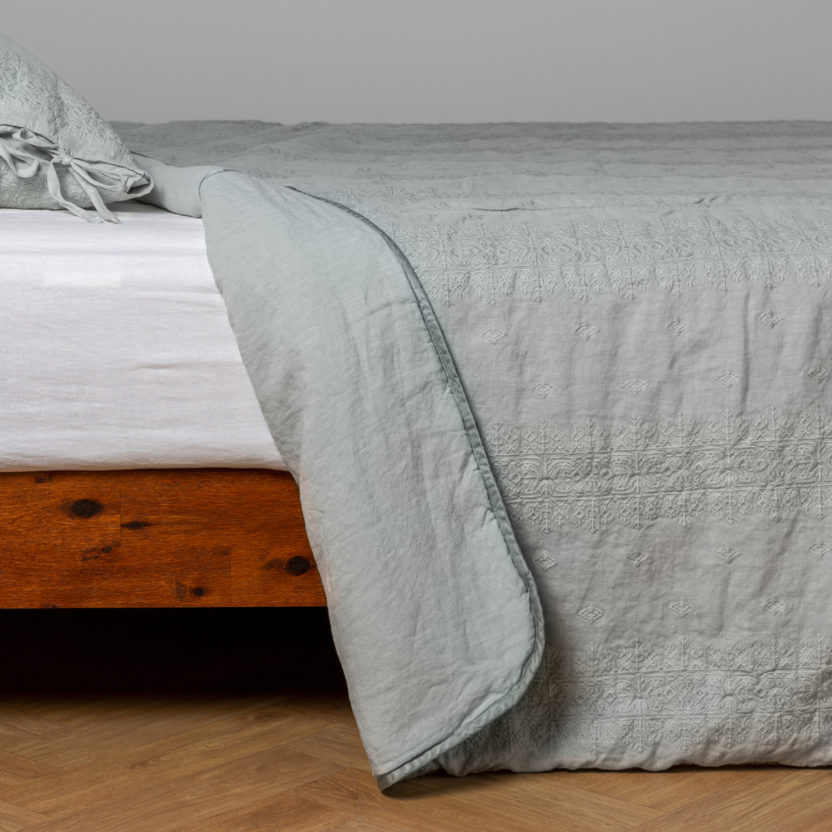 Eucalyptus: White: embroidered bedspread with corner pulled back showing the plain linen back. Shown with a white fitted sheet and matching sham, the bedframe is slightly visible and the bed is against a white wall and on a medium wood floor.
