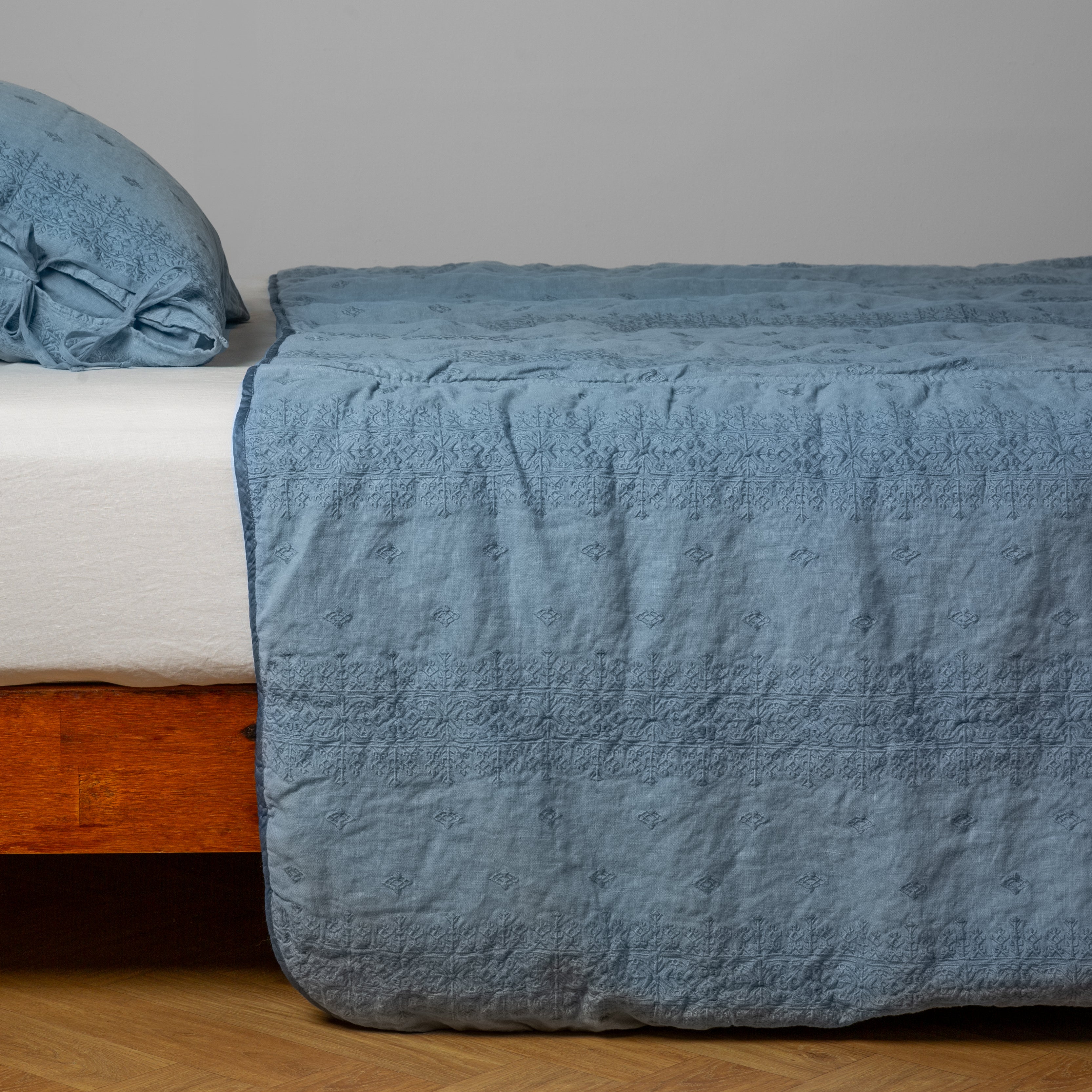 Tahoe Blue: embroidered midweight linen bedspread with matching sham on a bed with a white fitted sheet. Shot from the side view, the bedframe is slightly visible against a white background and medium wood floor.