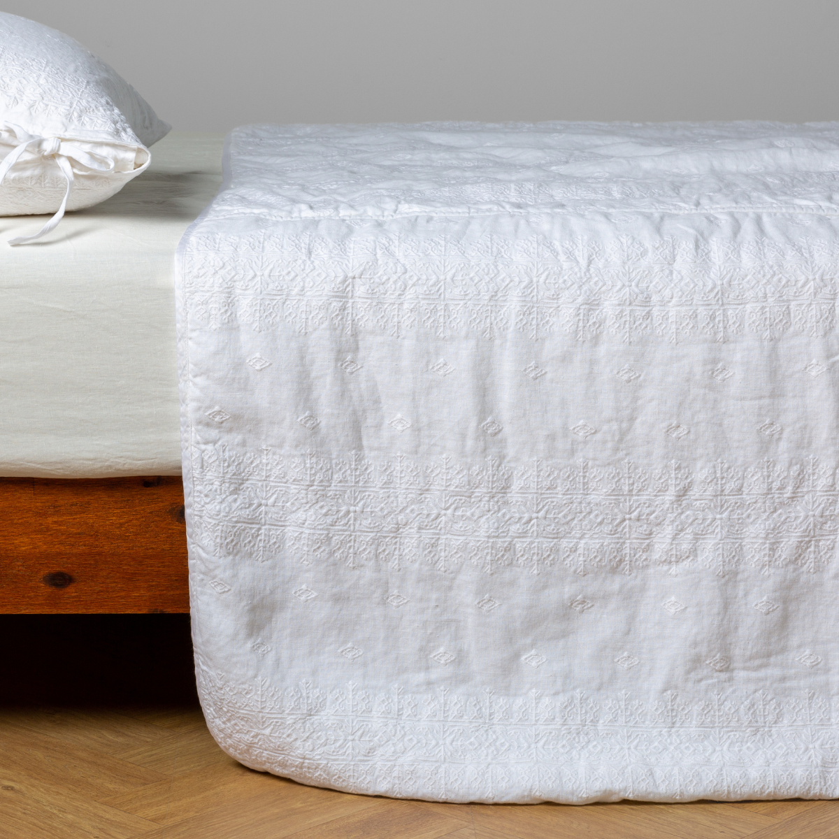 White: embroidered midweight linen bedspread with matching sham on a bed with a white fitted sheet. Shot from the side view, the bedframe is slightly visible against a white background and medium wood floor.