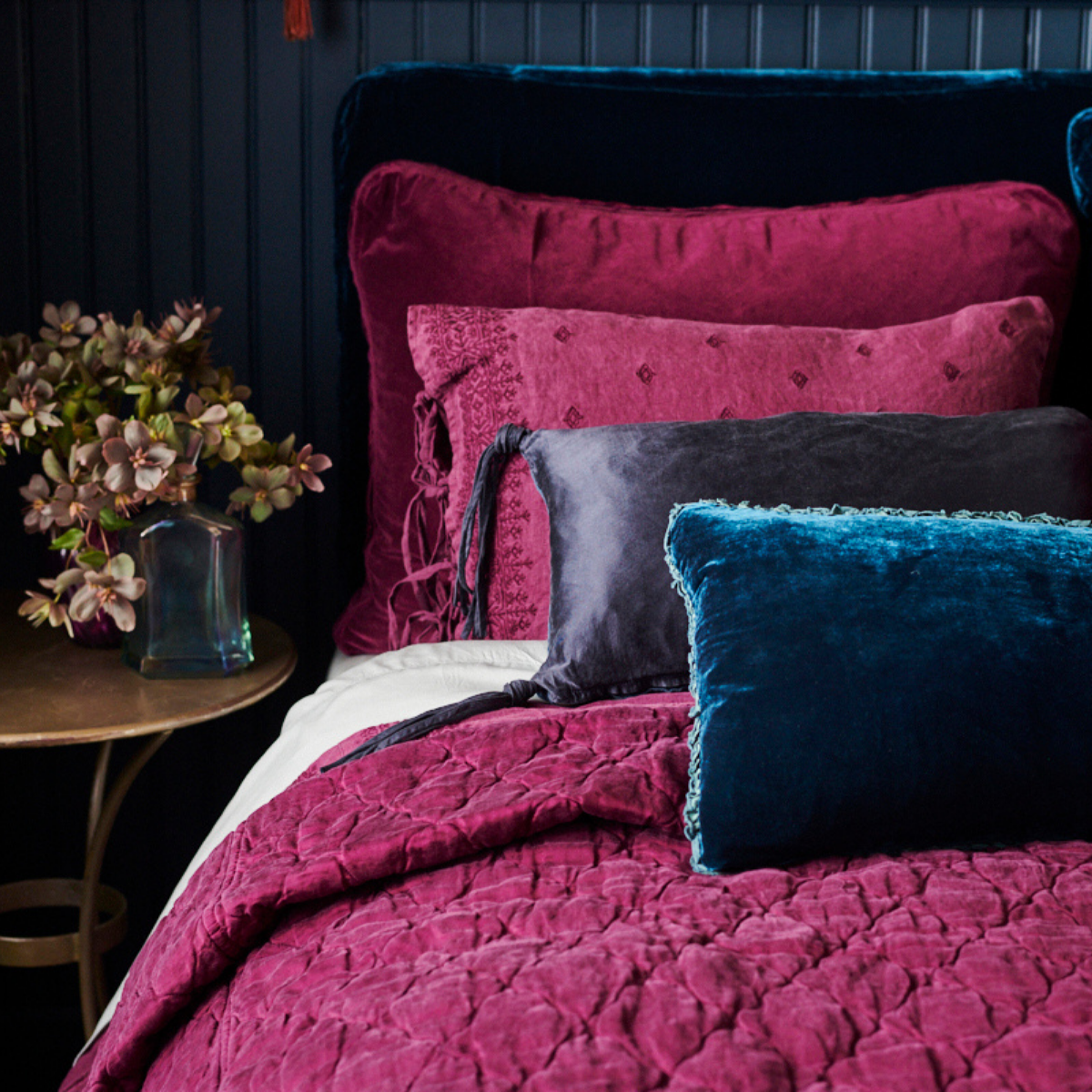 : Luna coverlet on a fig bed, with mixed textures of cotton velvet, embroidered linen, tassled silk charmeuse, and silk velvet. Deep blue throw pillows and dark blue background create a moody vibe.