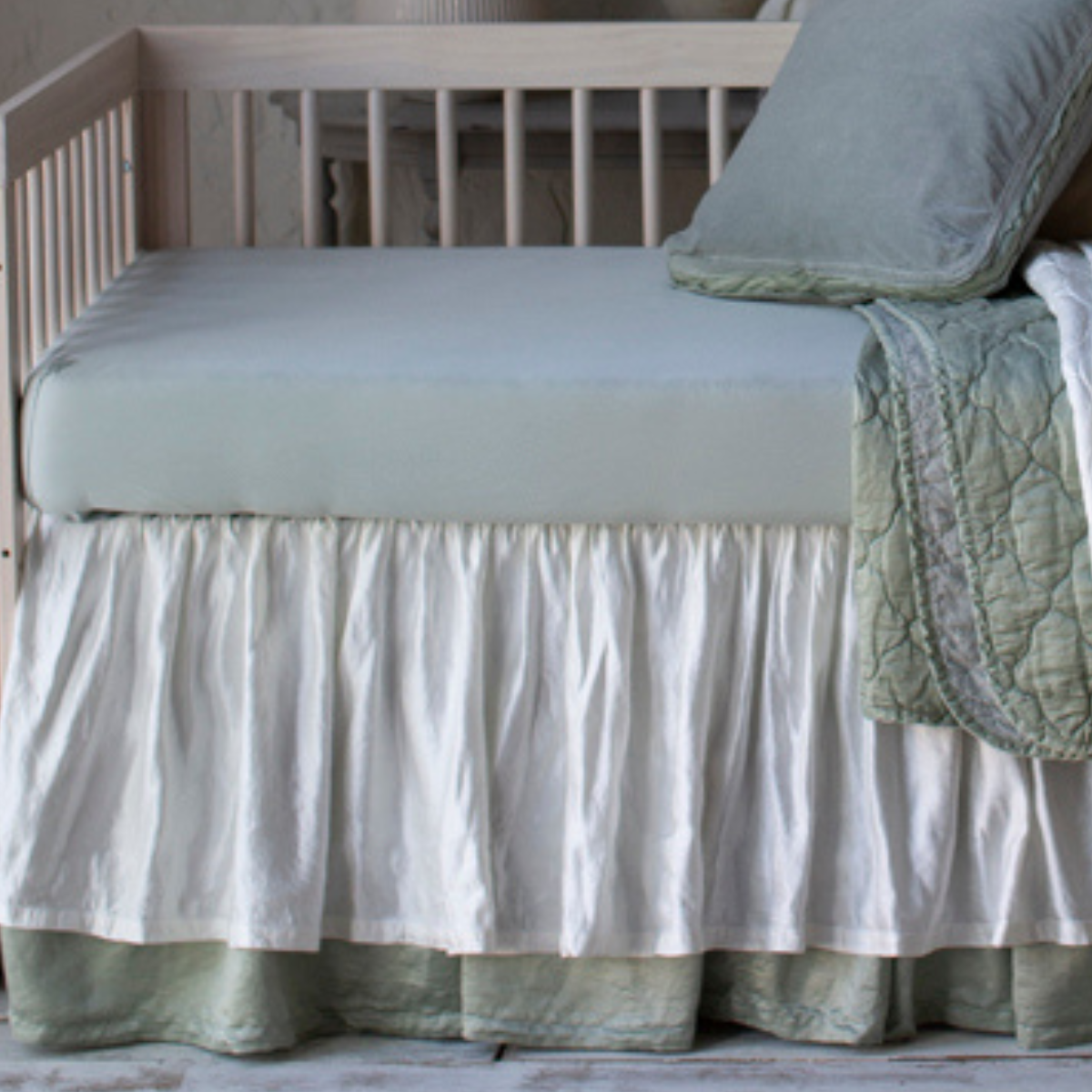 : A close up of crib sheet in eucalyptus shown in a soft green and cream toned nursery with varied textures.