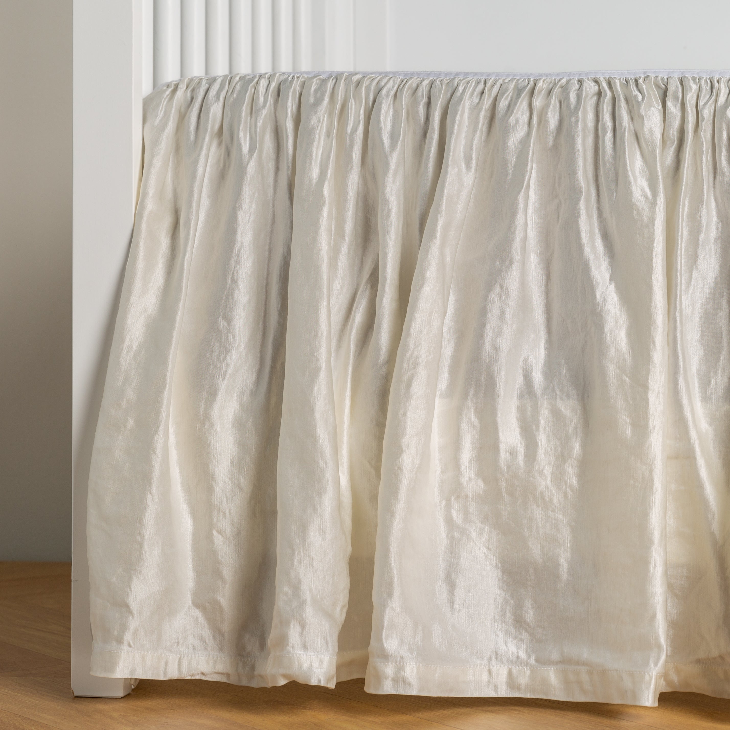 Winter White: charmeuse crib skirt shown on a white crib against a white wall and medium wood floor.