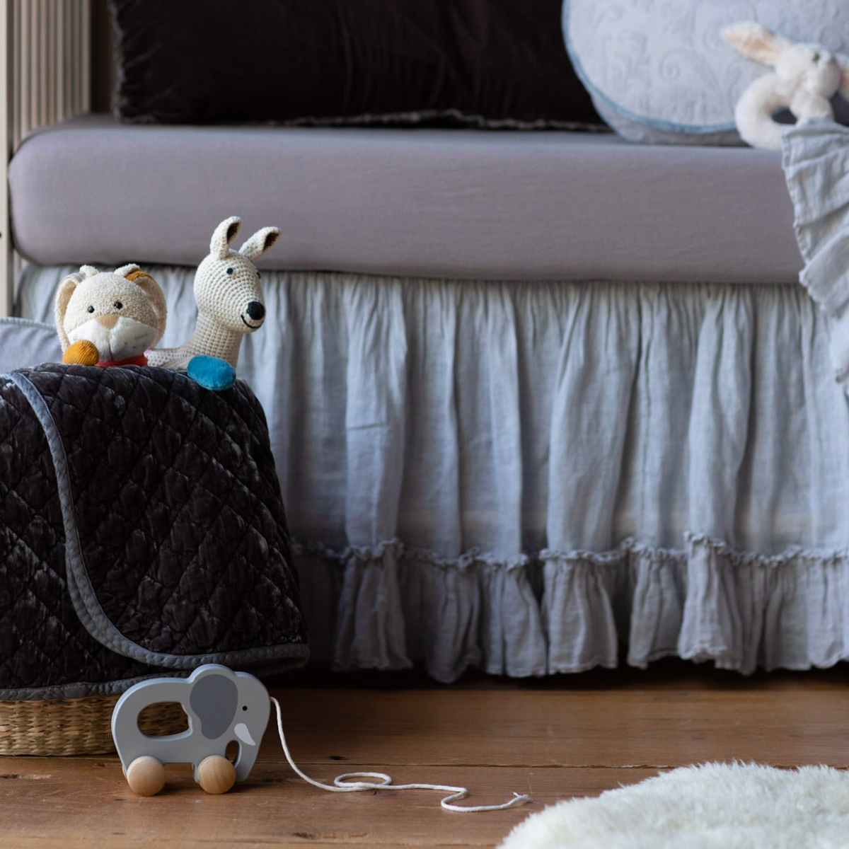 : Blanket in moonlight folded and draped over a basket in blue and grey nursery scene, shown with stuffed animals and wooden elephant pull toy.