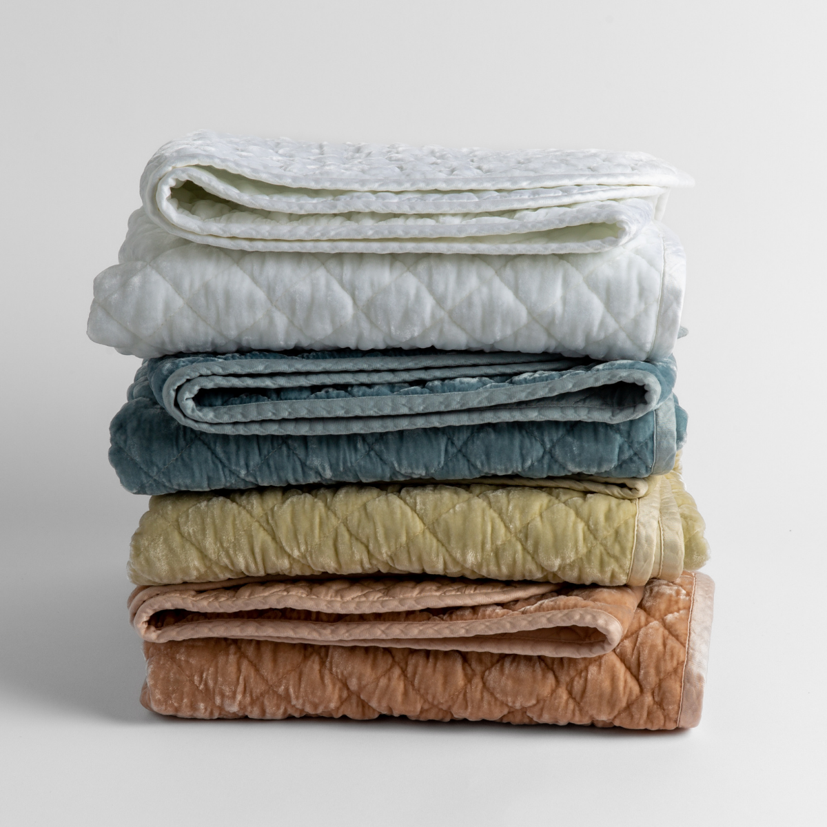 : stack of 4 quilted silk velvet baby blankets in white, cloud, parchment and rouge against a white background.