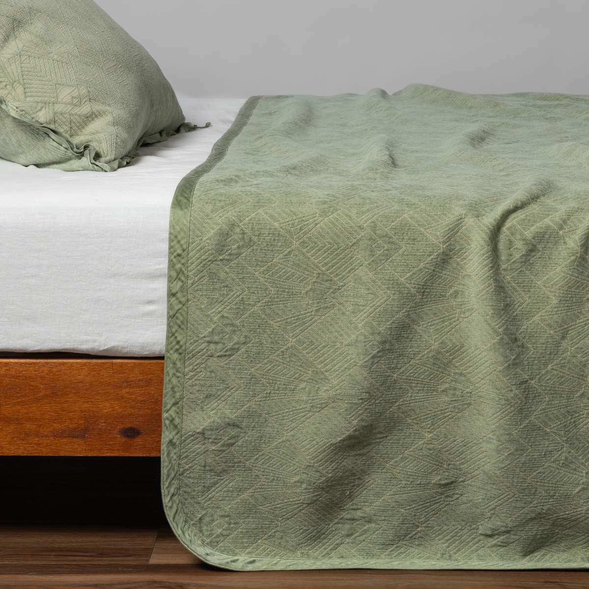 Desert Sage: matching trettino bedspread and sham in desert sage, a serenely elegant green, offering a hushed hue, shown from the side on a bed against a plain background.