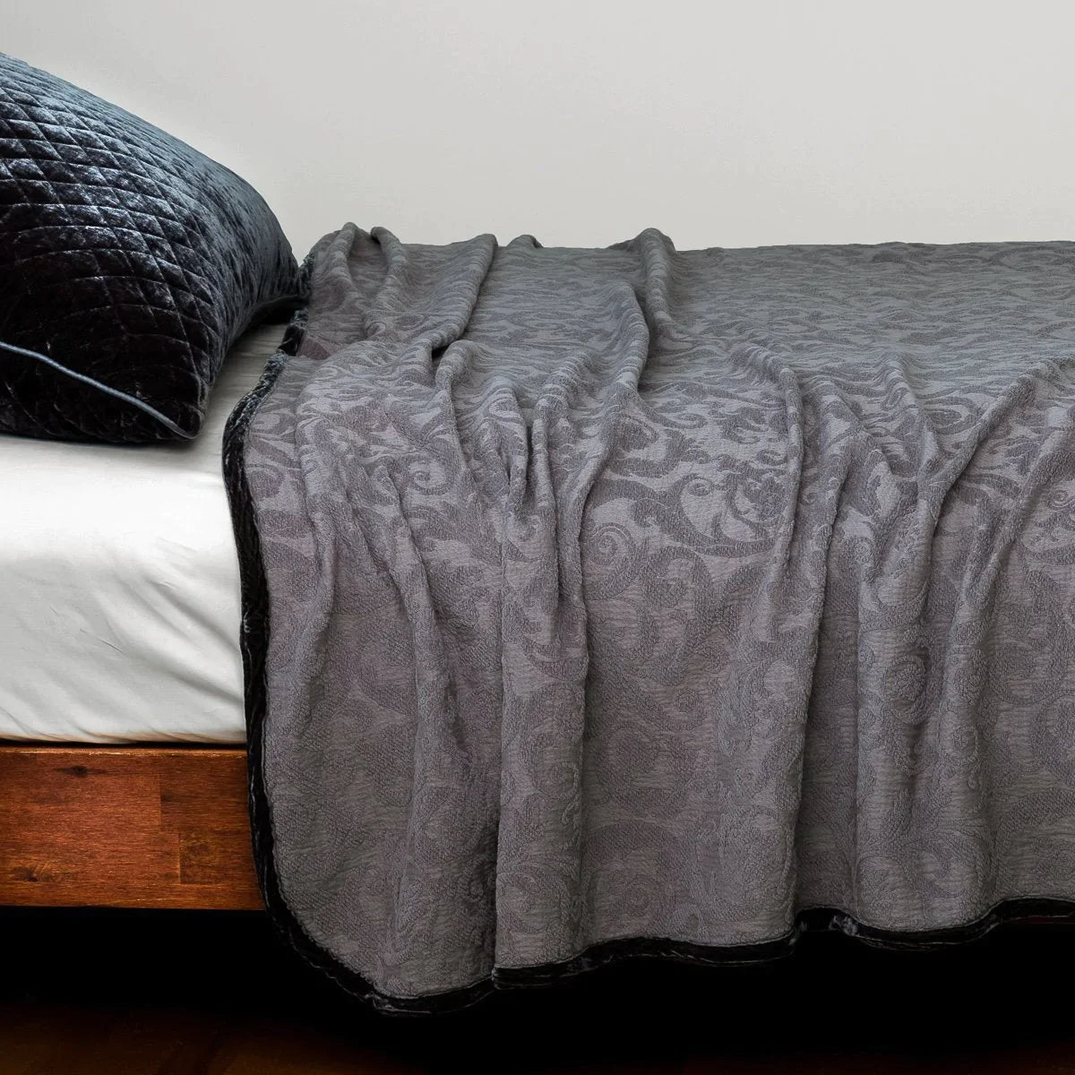 : The Adele coverlet in moonlight, styled on a wood framed bed shown from the side. Shown with a white sheet and a silk velvet quilted sham in moonlight.