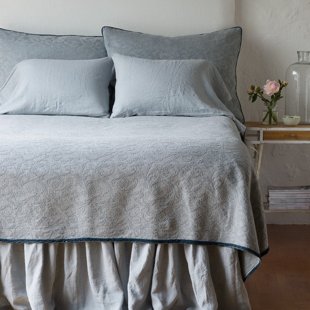 Mineral: The Adele coverlet in mineral, viewed from the foot of the bed, with white bed skirt and pillows visible. The headboard is plain light color, and part of a bedside table is visible with flowers, a vase and some books. against a plain white wall.