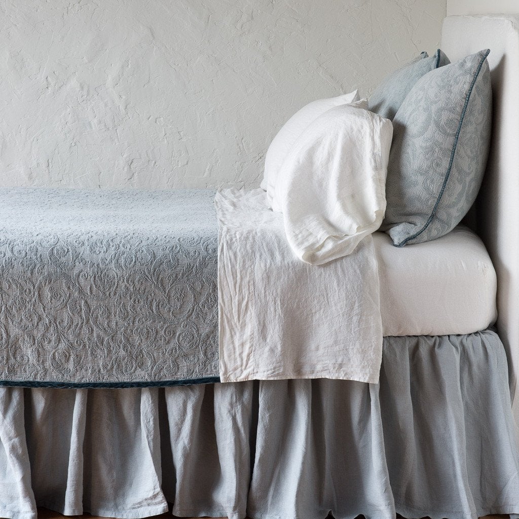 Mineral: The Adele coverlet in mineral, on a bed viewed from the side against a plain white wall. The bed is styled neatly with the coverlet folded back to reveal white sheets, bed skirt and pillows.