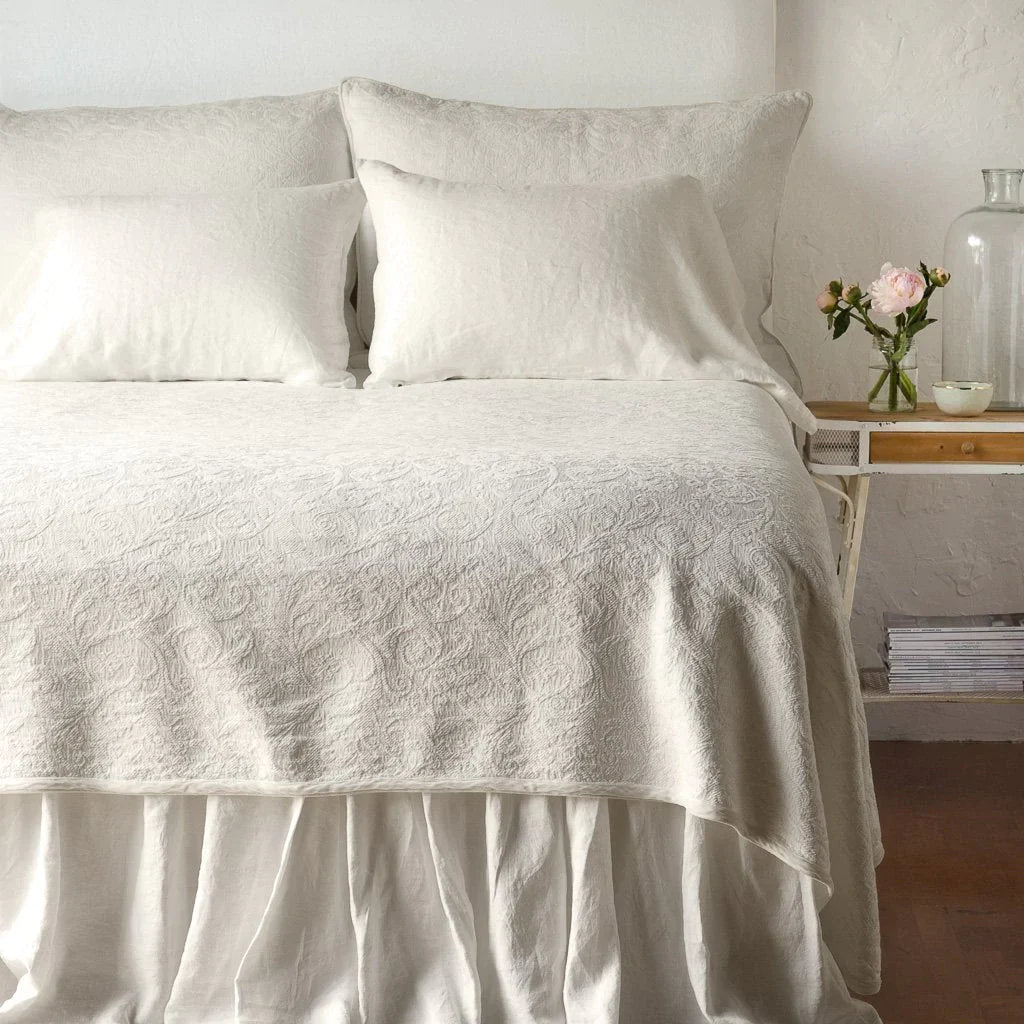 Parchment: The Adele coverlet in parchment, viewed from the foot of the bed, with parchment bed skirt and pillows visible. The headboard is plain light color, and part of a bedside table is visible with flowers, a vase and some books. against a plain white wall.
