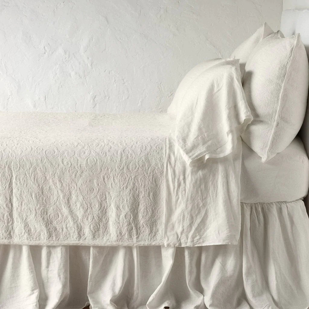 Parchment: The Adele coverlet in parchment, on a bed viewed from the side against a plain white wall. The bed is styled neatly with the coverlet folded back to reveal parchment sheets, bed skirt and pillows.