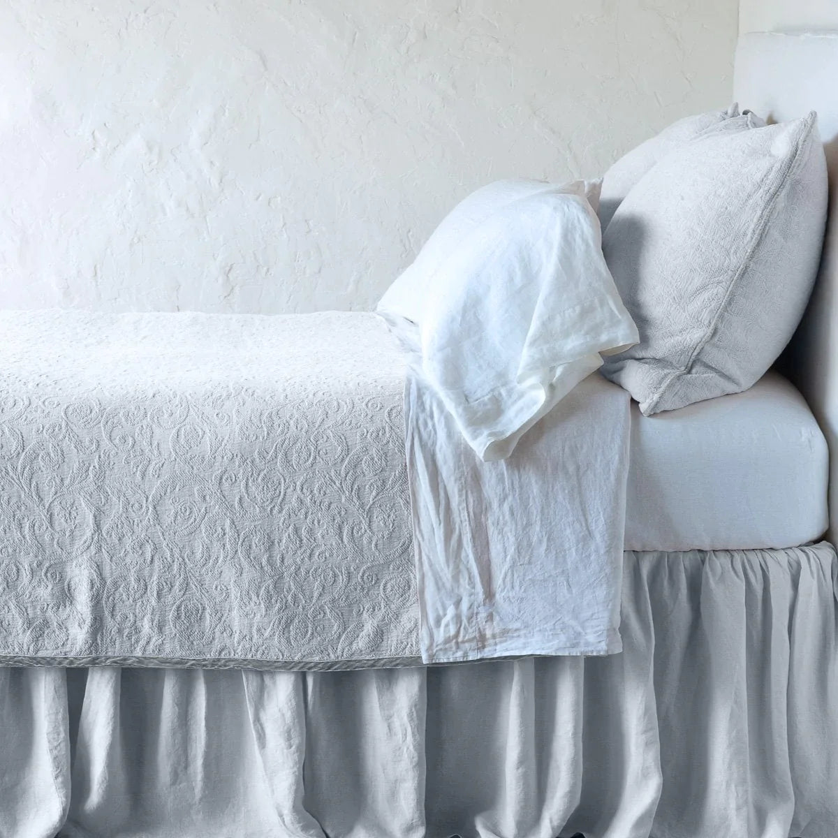 Sterling: The Adele coverlet in sterling, on a bed viewed from the side against a plain white wall. The bed is styled neatly with the coverlet folded back to reveal white sheets, bed skirt and pillows.