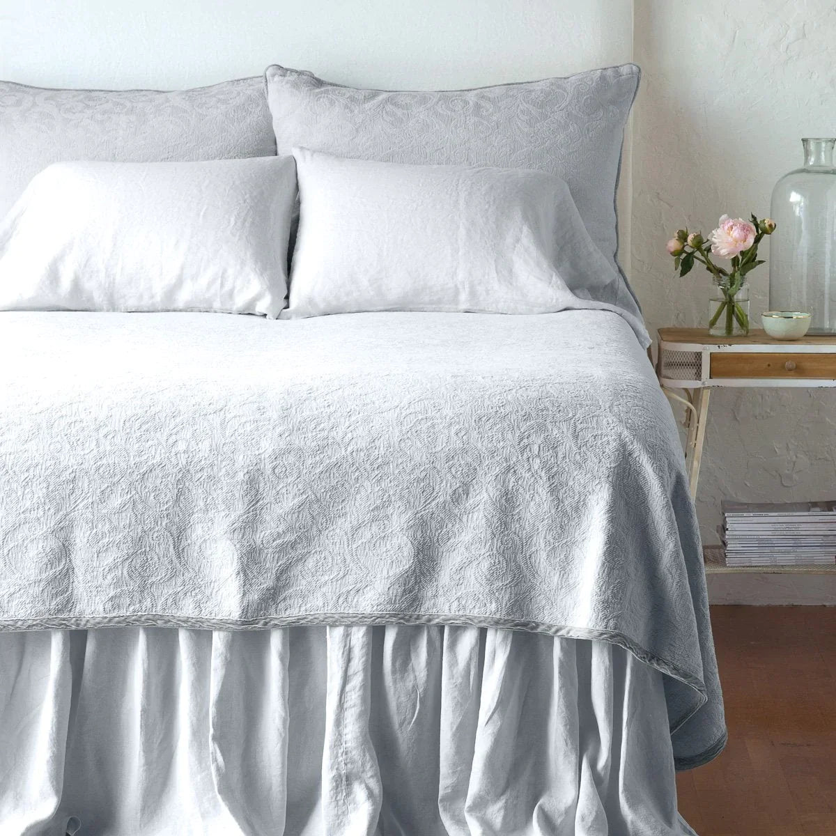 Sterling: The Adele coverlet in sterling, viewed from the foot of the bed, with white bed skirt and pillows visible. The headboard is plain light color, and part of a bedside table is visible with flowers, a vase and some books. against a plain white wall.