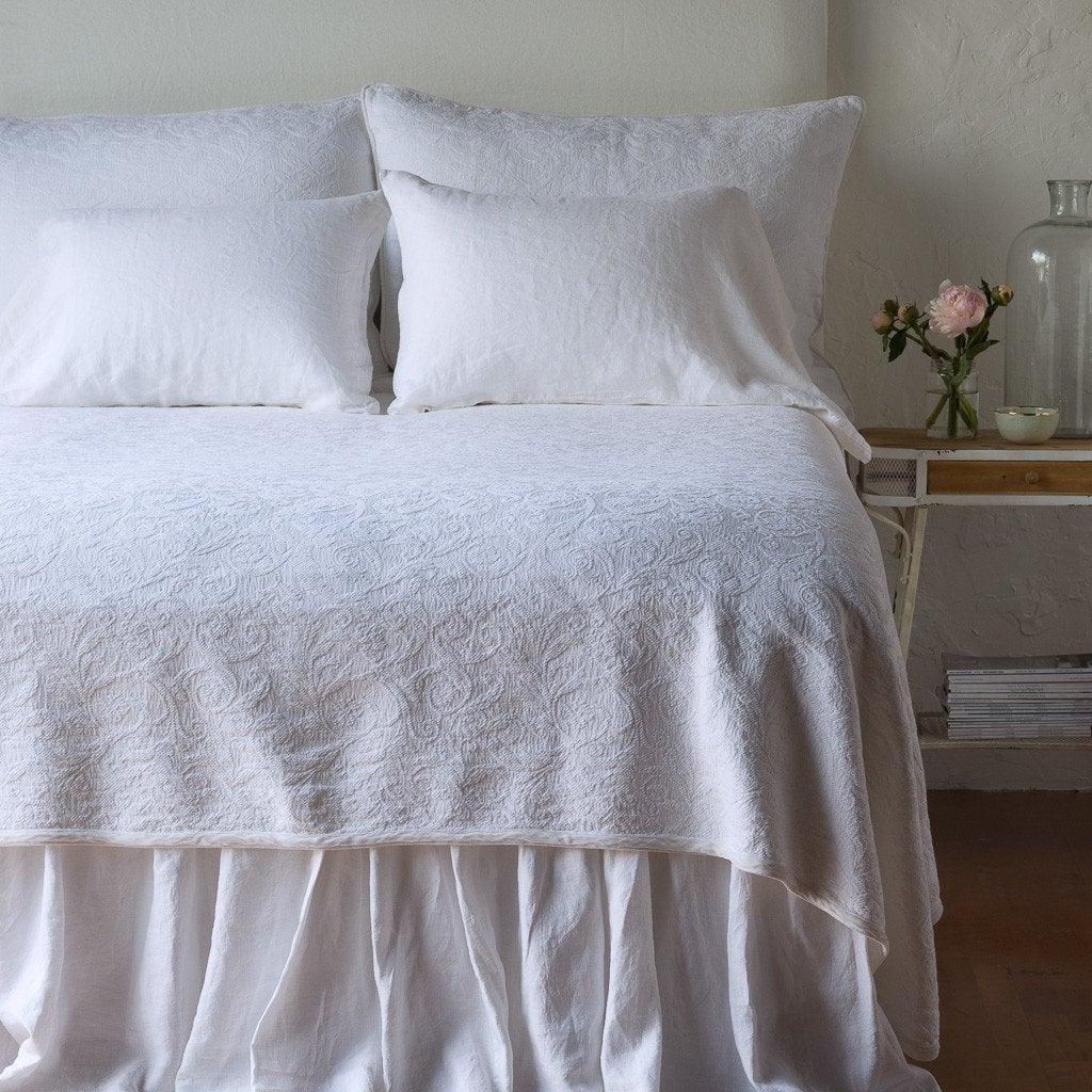 White: The Adele coverlet in white, viewed from the foot of the bed, with white bed skirt and pillows visible. The headboard is plain light color, and part of a bedside table is visible with flowers, a vase and some books. against a plain white wall.
