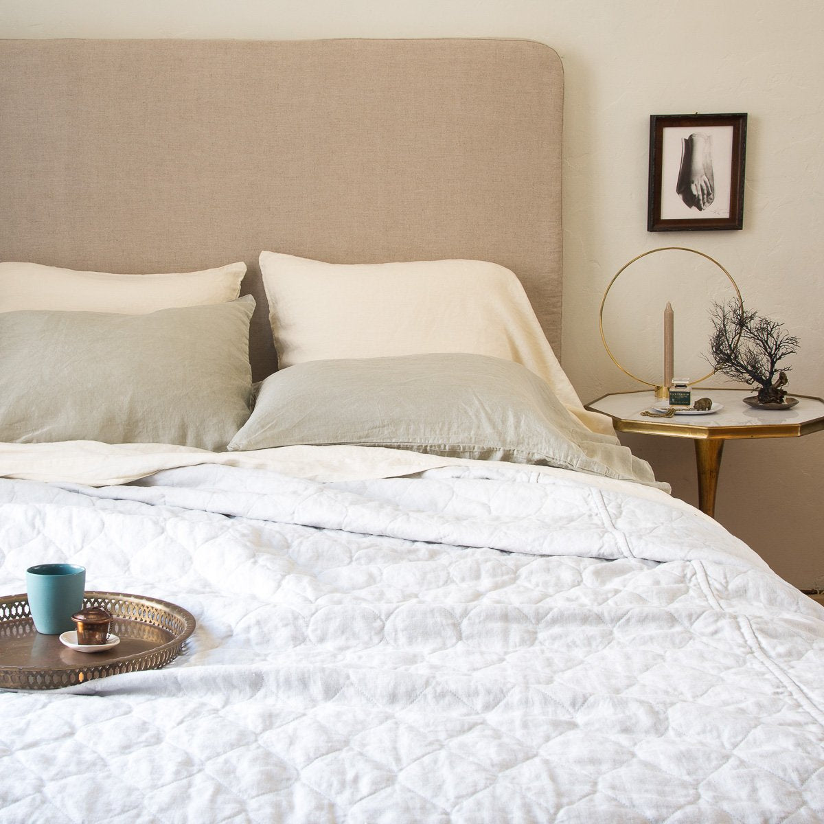 : White Austin quilted coverlet shown partially unmade from the foot of the bed with light neurtal sheets and pillowcases, against a beige headboard. Styled with a tray on the bed and simple bedside table and art.