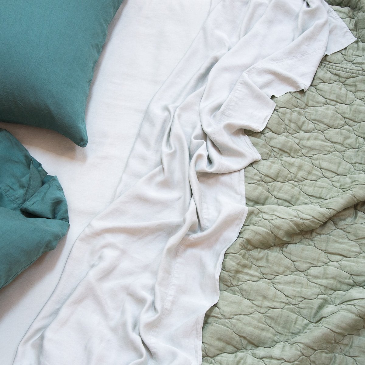 : Luna coverlet in eucalyptus on a rumpled bed with white sheets and jade sleeping pillows - overhead view.