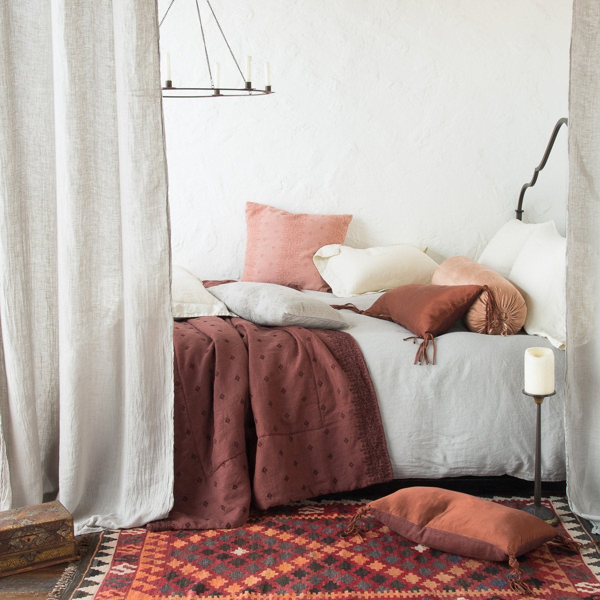 : Ines throw blanket in mahogany, layered on a rumpled, blue, brown, and pink toned bed in linen and charmeuse, with a geometric rug and wrought-iron room accents - side view.