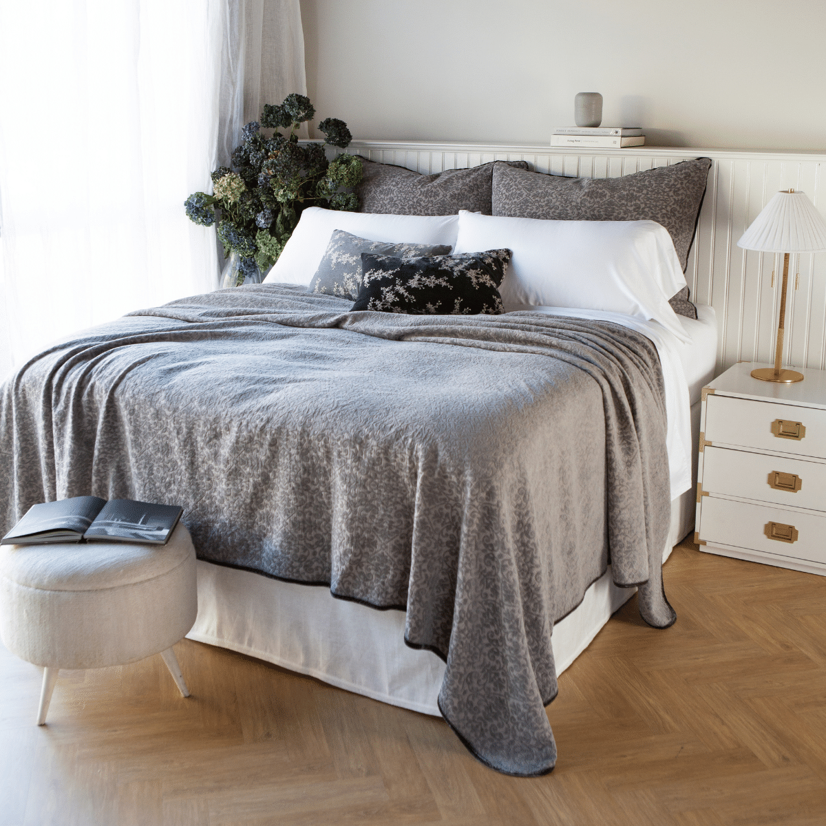 : Vienna coverlet and shams in moonlight grey on a simple, elegant bed with white cotton sheeting. Three-quarter angle with herringbone wood floor and white furnishings with gold accents.