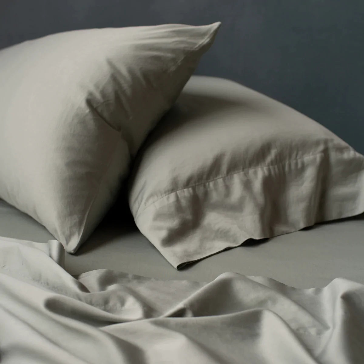 : Rumpled Bria flat sheet in eucalyptus in the foreground, with matching sleeping pillows angled asymmetrically against a moody dark background.