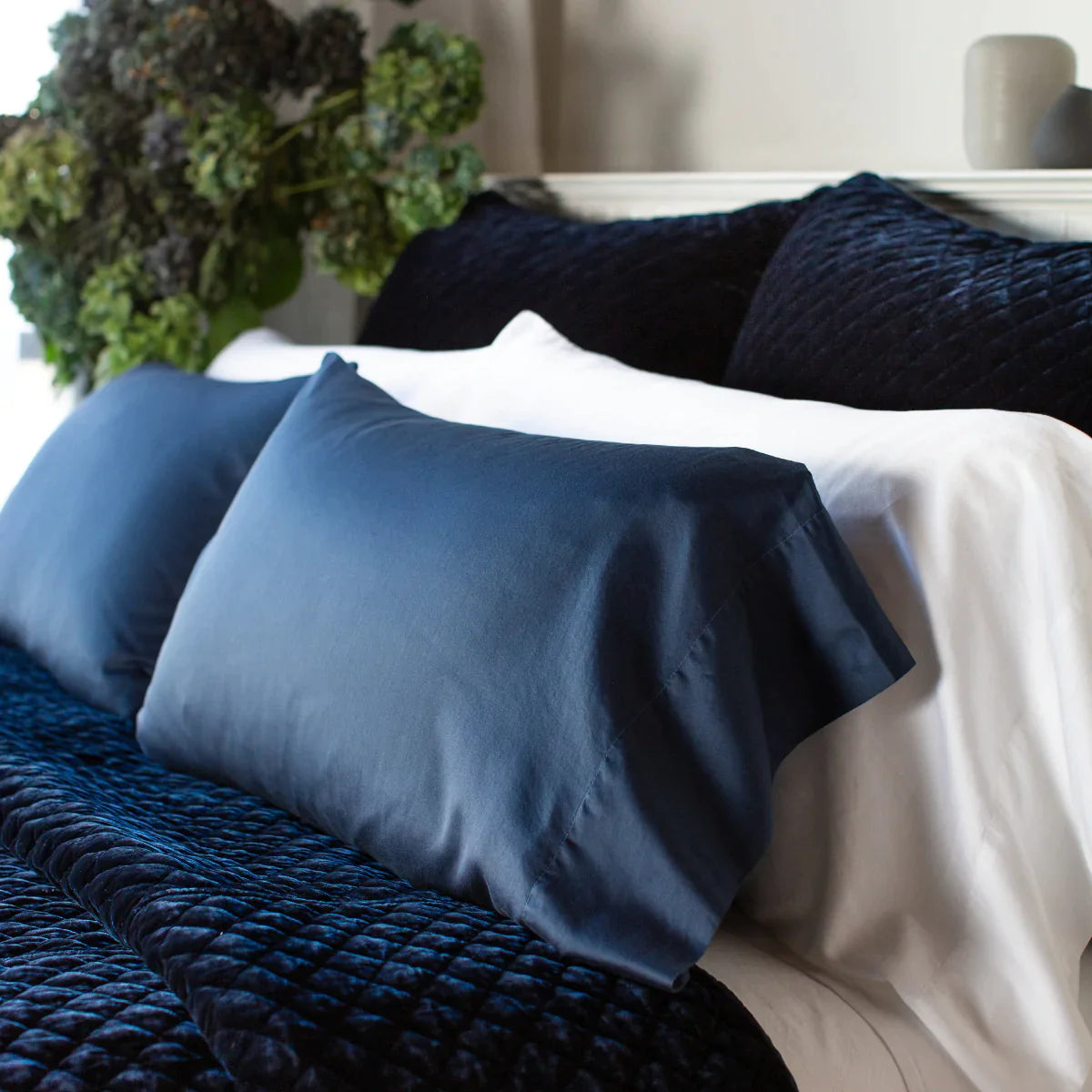 : Bria pillowcases in white and midnight shown from three quarter close up angle. The cotton sateen shines against the deep blue silk velvet quilted shams and coverlet.