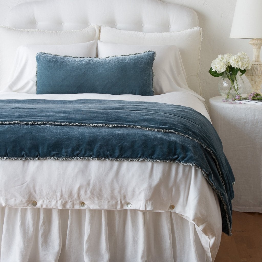 Mineral: Carmen 16x36 pillow and throw blanket in mineral, shown on an otherwise all white, neatly made bed - foot of bed angle.
