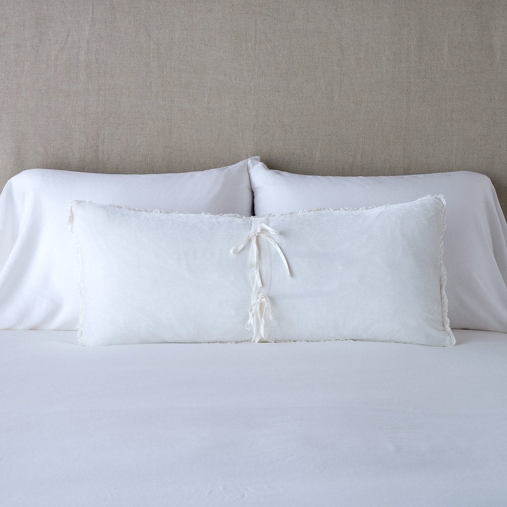 White: Silk velvet lumbar pillow with petite ruffle leaning upright and backwards against neutral bedding and headboard, showcaseing the silk charmeuse tie closures.