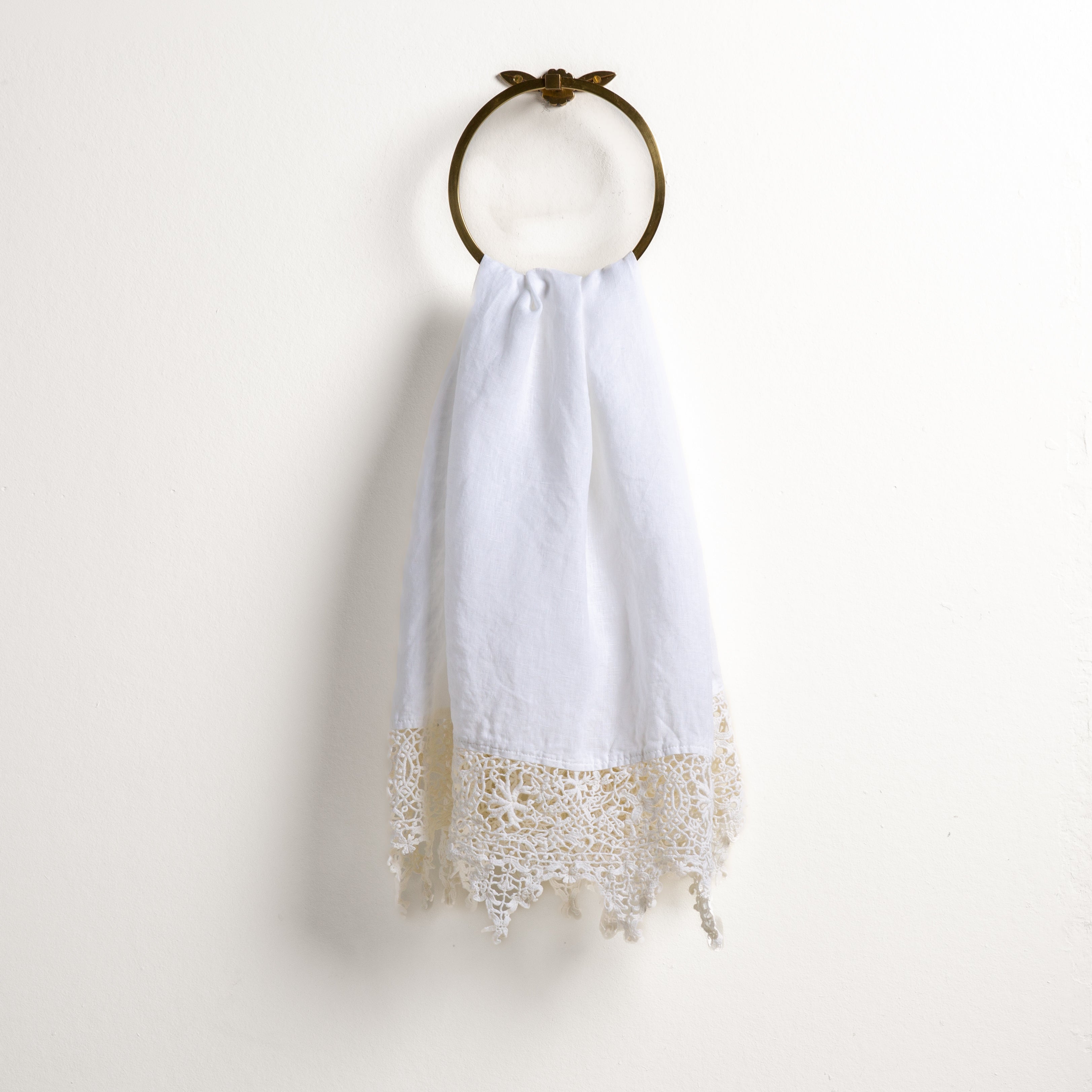 White: Lace trimmed linen guest towel draped through a decorative towel ring against a plain white background.