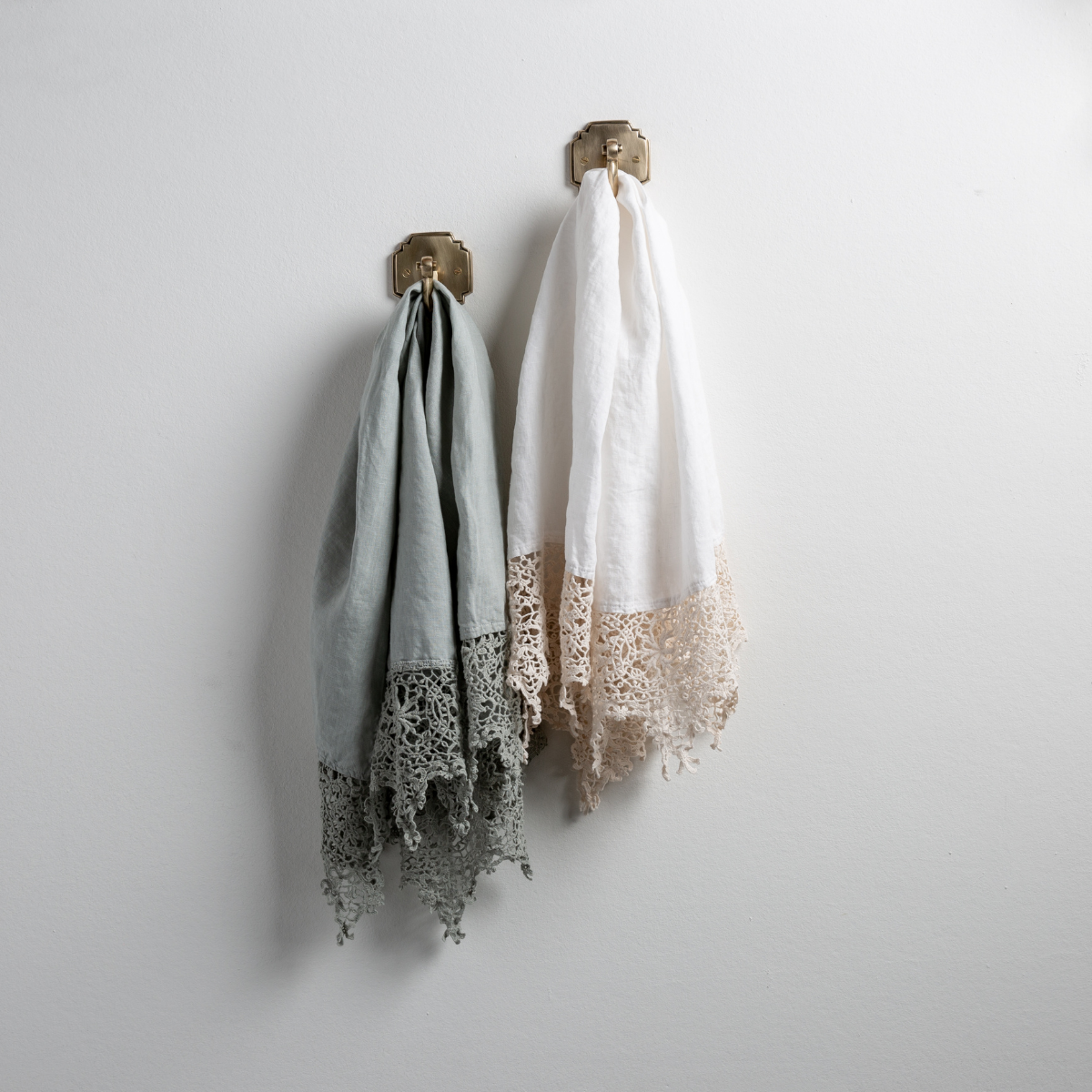 : Two Frida guest towels, draped over decorative hooks against a white wall - eucalyptus and winter white