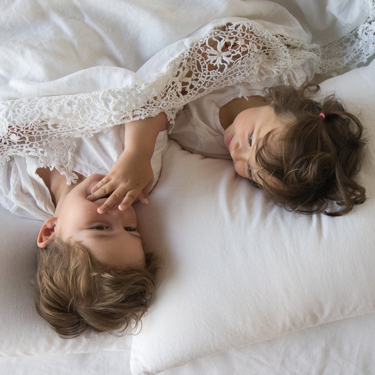 : Two young children laying in a white bed under a Frida flat sheet - overhead view.