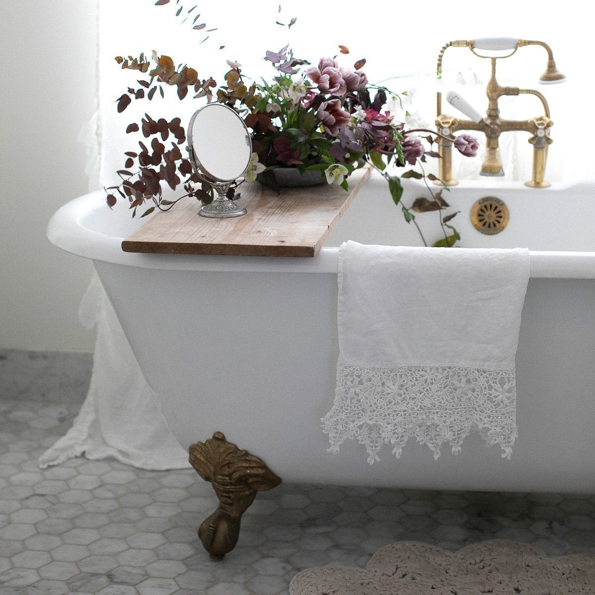 : White Frida guest towel draped over the edge of an antique tub, with copper accents and lavender and green floral bouquet.